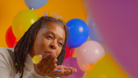 Studio-Portrait-Of-Woman-Wearing-Birthday-Headband-Celebrating-Blowing-Paper-Party-Confetti